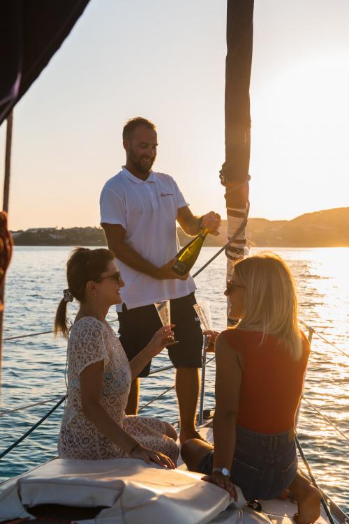 Skipper versa vino bianco a due ragazze durante aperitivo in barca a vela a Stintino